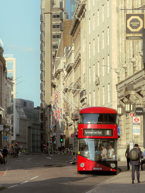 A busy urban street scene with a modern double-decker red bus on the road, surrounded by classical-style buildings in cream and beige tones. The street is lined with commercial signage, including a bl